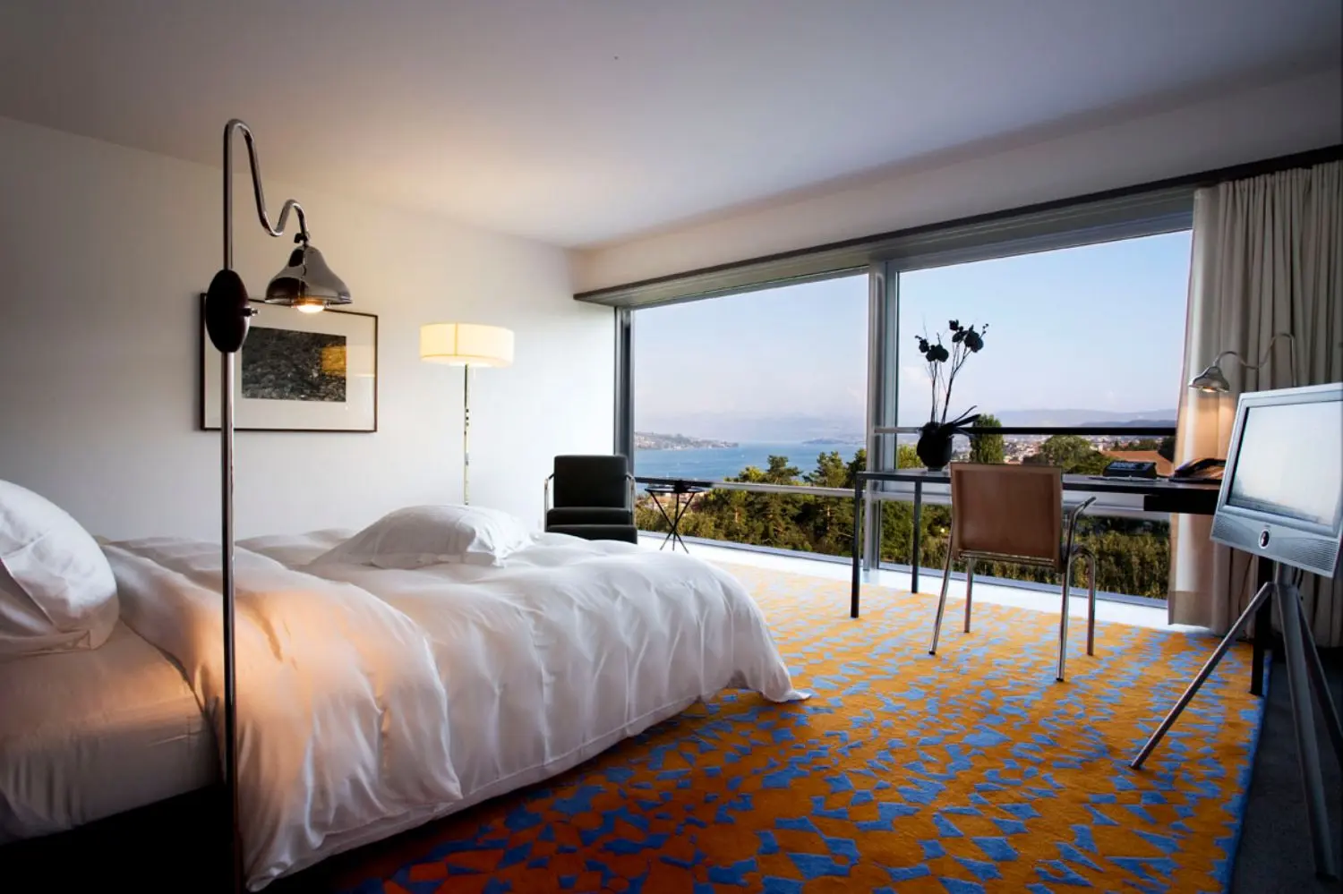 A bedroom with a bed, light, desk and chair with windows overlooking lake Zurich.