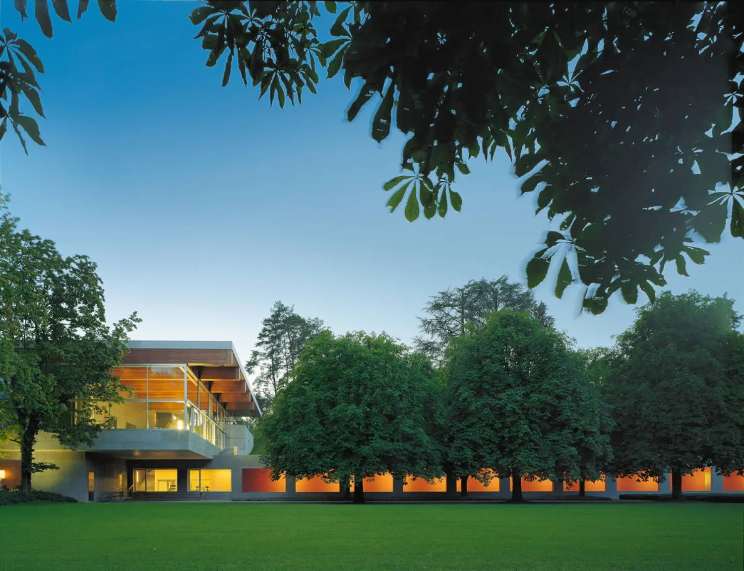 Orange tinged lights in a concrete building and hallway extension among green grass and trees with a blue sky.
