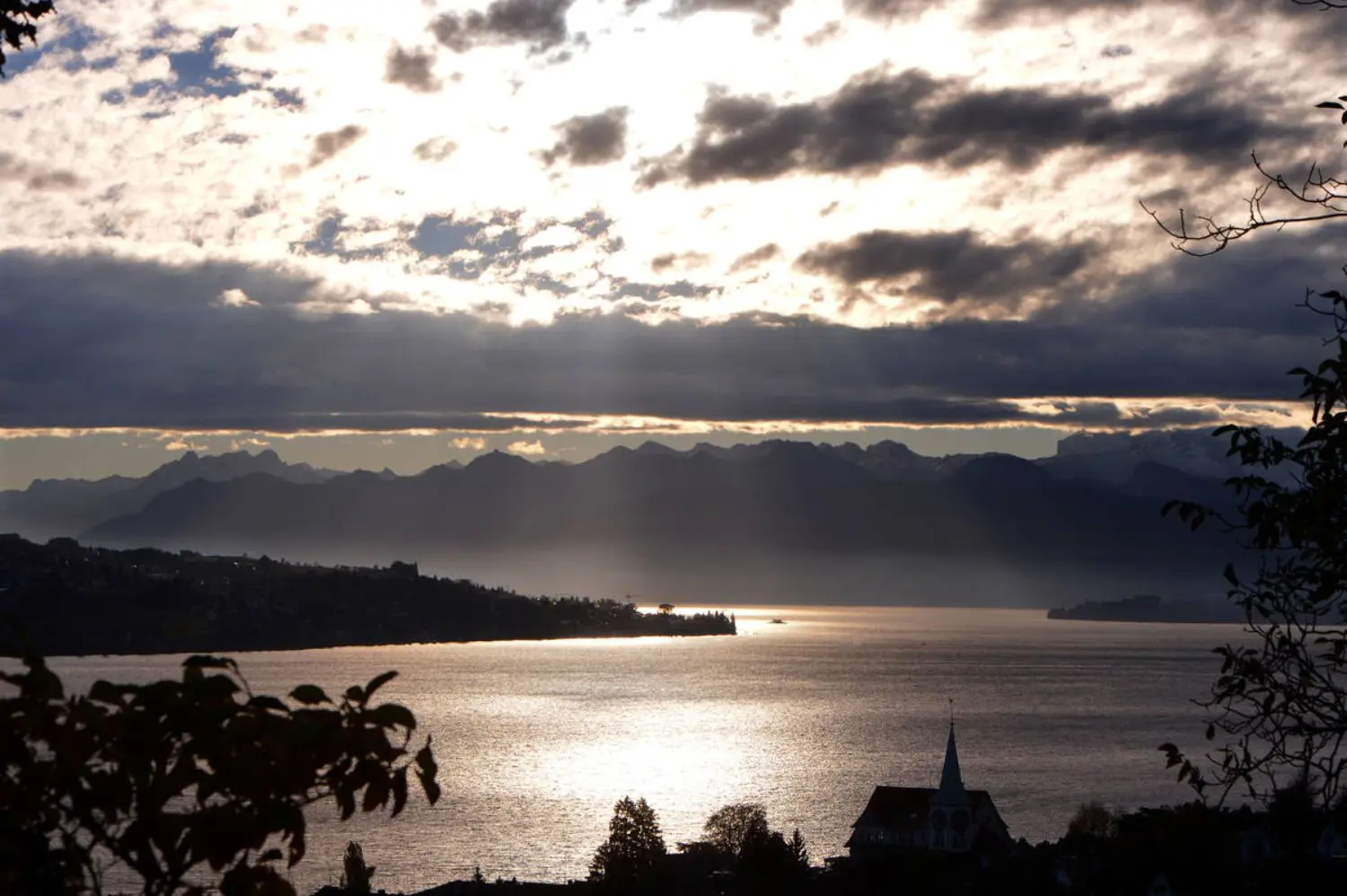 A shiny lake with a mountain range in the background and clouds in front of the sun.