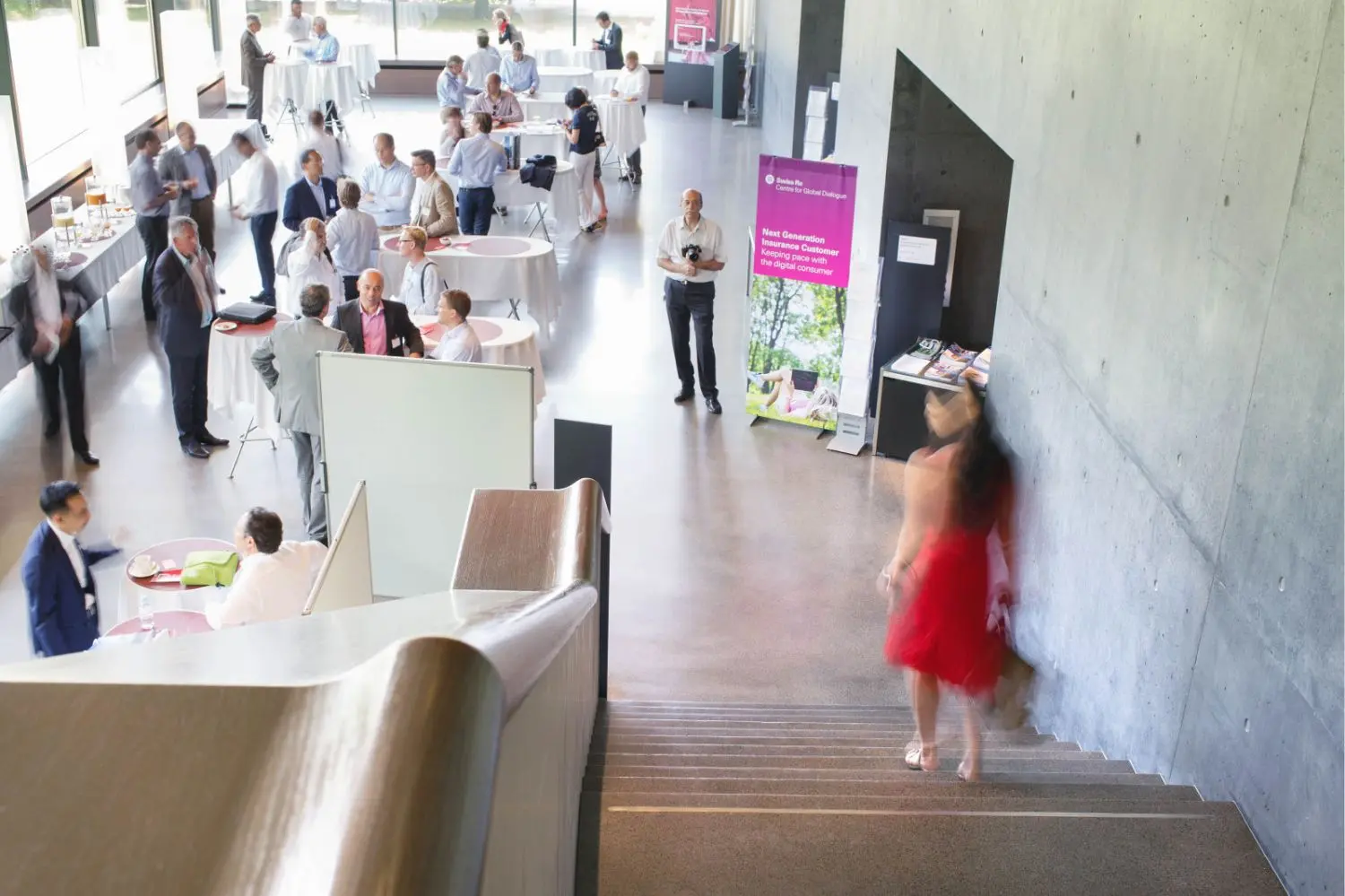 A woman in a red dress descending stairs to a lobby with tables and people.